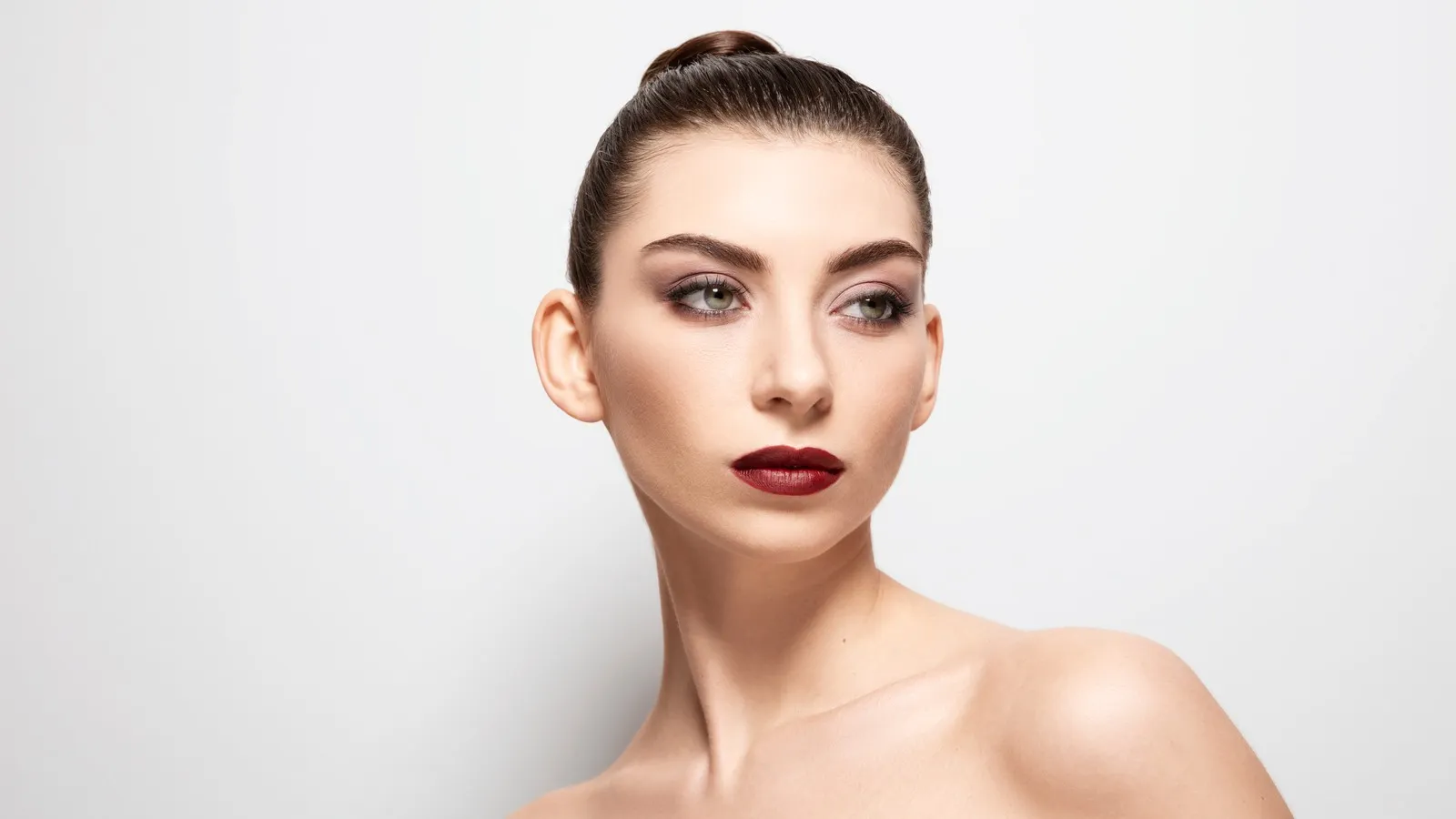 Eine Frau mit strengem Zopf schaut in die Ferne. Ihr Make-up vereint verschiedene Bronze-Töne. Die Lippen sind in einem dunklen Rotbraun geschminkt.